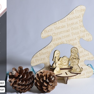 Op de afbeelding: Een houten kerstboomuitsparing met een kerststal erin. De boom is versierd met de woorden "Vrolijk Kerstfeest" in meerdere talen. De kerststal bevat Maria, Jozef, baby Jezus, een ezel en een koe.