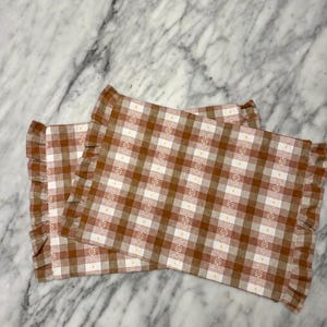 May include: Two rectangular placemats featuring a brown and white gingham check pattern with small white floral accents. Each placemat is finished with a gathered ruffle trim along the two shorter sides, creating a textured edge detail.