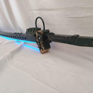 Puede incluir: Una espada larga de color verde oscuro con un diseño mecánico, que presenta una luz azul brillante a lo largo del filo de la hoja. La empuñadura incluye un bucle circular, detalles en oro metálico y complejos elementos estructurales.
