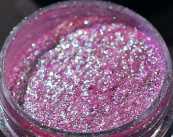 May include: A small jar of pink glitter with iridescent shimmer. The jar has a clear lid and a black base with a logo.
