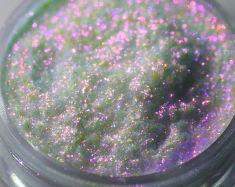 May include: A small jar of iridescent green glitter with a white base. The glitter has a holographic finish and reflects light in a variety of colors.