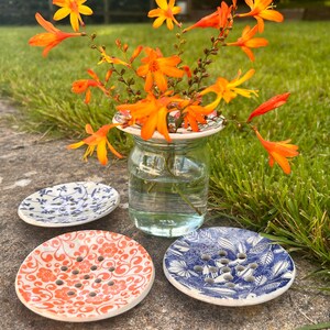 May include: A glass jar with orange flowers and three ceramic soap dishes. The dishes have floral and leaf patterns in blue and orange. The flowers are a vibrant orange colour. The dishes are round.