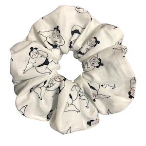 May include: White scrunchie featuring a repeating pattern of sumo wrestlers in black and pink. The fabric is gathered and elasticated, creating a voluminous hair accessory. The sumo wrestlers are depicted in various poses.