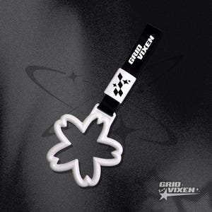 May include: A white, flower-shaped accessory with a black strap. The strap features the text "GRID VIXEN" in white, along with a geometric design. The accessory is set against a dark, textured background.