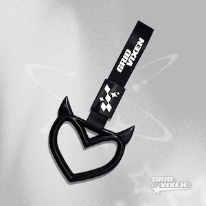 May include: Black heart-shaped car accessory with devil horn accents. The accessory has a black strap with the text "GRID VIXEN" in white. A white and black logo is on the strap.