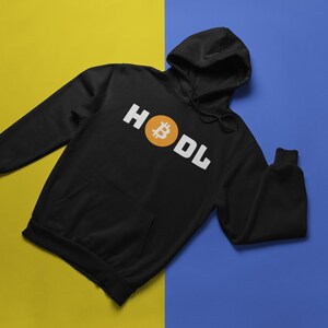 以下が含まれることがあります： 白い文字で「HODL」という言葉とオレンジ色のビットコインのシンボルが入った黒いパーカー。 パーカーにはフロントポケットとドローストリングフードが付いています。 背景は黄色と青に分かれています。