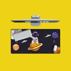 May include: A black and white mousepad with a cartoon astronaut sitting on a planet. The astronaut is wearing a white spacesuit and a helmet. The planet is surrounded by stars and a crescent moon. The mousepad also features a keyboard and a spaceship.