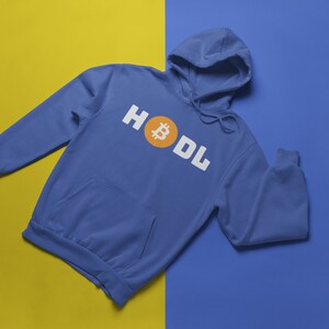 以下が含まれることがあります： 白いブロック文字で「HODL」という言葉とオレンジ色のビットコインのシンボルが入った青いパーカー。 パーカーにはフロントポケットとフードが付いています。 背景は黄色と青に分かれています。