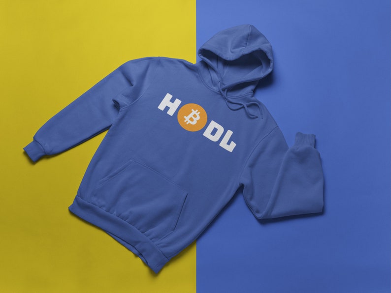 以下が含まれることがあります： 白いブロック文字で「HODL」という言葉とオレンジ色のビットコインのシンボルが入った青いパーカー。 パーカーにはフロントポケットとフードが付いています。 背景は黄色と青に分かれています。