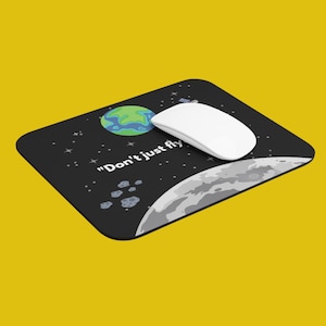 Może przedstawiać: Czarna podkładka pod mysz z motywem kosmicznym. Zawiera kreskówkową grafikę Ziemi, Księżyca i gwiazd. Na podkładce pod mysz nadrukowany jest tekst "Don't just fly".