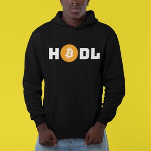 以下が含まれることがあります： 黒いパーカー。白い文字で「HODL」と書かれています。「O」は金のビットコインのシンボルに置き換えられています。パーカーにはフロントポケットとフードが付いています。背景は黄色です。