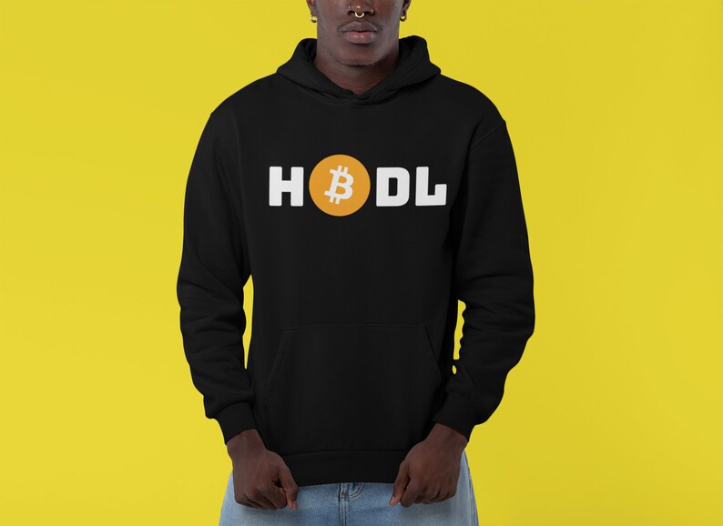 以下が含まれることがあります： 黒いパーカー。白い文字で「HODL」と書かれています。「O」は金のビットコインのシンボルに置き換えられています。パーカーにはフロントポケットとフードが付いています。背景は黄色です。