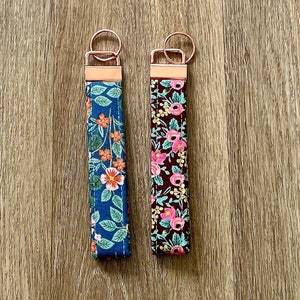 May include: Two floral patterned keychains with rose gold key rings. The keychain on the left has a blue background with pink and orange flowers. The keychain on the right has a maroon background with pink and yellow flowers.
