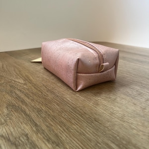 May include: A small, rectangular, pink cosmetic bag with a zipper closure. The bag is made of a textured material and sits on a wooden surface. The zipper is a matching rose gold color.