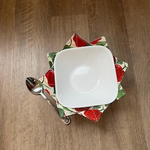 May include: A white ceramic bowl with a red and green apple print bowl cozy. The bowl cozy is shaped like a star and has a white background with red apples and green leaves.