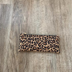 May include: A rectangular pencil case with a leopard print pattern. The case is primarily tan with dark brown spots, and features a gold-colored zipper closure.