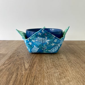 May include: A blue bowl cozy with a sea turtle pattern. The cozy is made of fabric and has a star-shaped design. The cozy is sitting on a wooden surface.