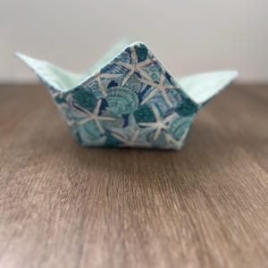 May include: A light blue fabric bowl with a seashell and starfish pattern. The bowl is folded into a star shape and is sitting on a wooden surface.