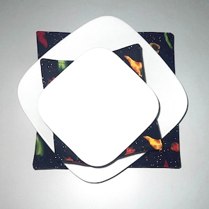 May include: A stack of three white square plates atop a square fabric placemat. The placemat has a dark blue background with a pattern of colorful peppers and stars. The plates are arranged in a nested fashion.