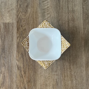May include: A white ceramic bowl with a rounded bottom sits on a pink and gold leopard print fabric coaster.