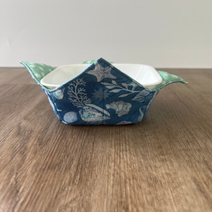 May include: A white ceramic bowl with a blue and green fabric bowl cozy. The cozy has a sea life pattern with white starfish, seashells, and seaweed.