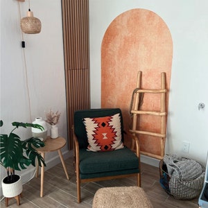 May include: A green armchair with a patterned pillow sits in a room with a wooden accent wall and a terracotta arch sticker on the wall. A wooden ladder leans against the wall and a woven basket sits on the floor.