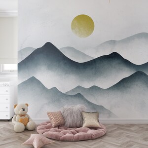 May include: A plush pink cushion with a fluffy white pillow and two gold pillows sits in front of a wall mural depicting a watercolor-style mountain range with a yellow sun.