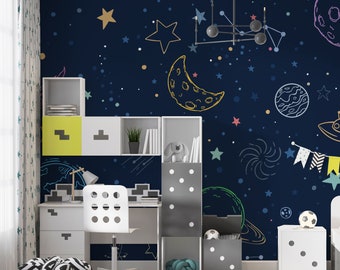 Puede incluir: Un dormitorio infantil con una pared azul marino que presenta un mural de temática espacial dibujado a mano. El mural incluye estrellas, planetas, una luna creciente y una galaxia. La habitación está decorada con un escritorio blanco, una silla blanca y una unidad de almacenamiento blanca con acentos amarillos.
