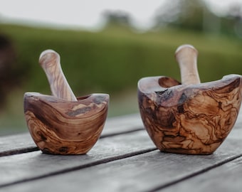 Könnte beinhalten: Zwei Mörser und Stößel aus Olivenholz. Die Mörser sind braun mit einem natürlichen Holzmaserungsmuster. Die Stößel sind glatt und hellbraun.