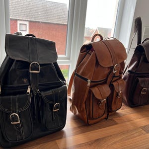 May include: Three leather backpacks in black, tan, and brown are displayed. Each bag has a flap closure with metal buckles, front pockets, and adjustable straps. The backpacks are designed for carrying items. The bags are on a wooden surface.