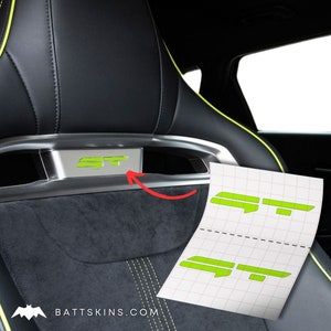 May include: A close-up of a car seat with a green "ST" emblem. A sheet of paper with two "ST" stencils is shown next to the seat.