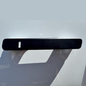 May include: A black, rectangular car door handle with a small, rectangular opening. The handle is mounted on a white surface, casting a shadow. The handle appears to be made of a matte material.