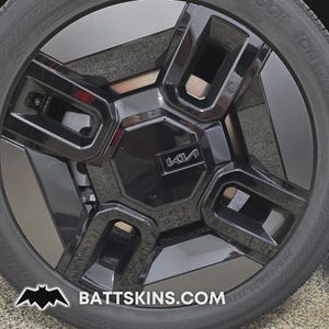 May include: Close-up of a black car wheel with a glossy finish. The wheel features a geometric design with the Kia logo in the center. The tire is black with white lettering. The website address battskins.com is visible at the bottom.
