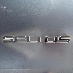 Puede incluir: Primer plano del exterior de un coche, que muestra el emblema cromado "SELTOS". Las letras están delineadas en plata, con un interior negro, sobre un fondo gris texturizado. El emblema está centrado.