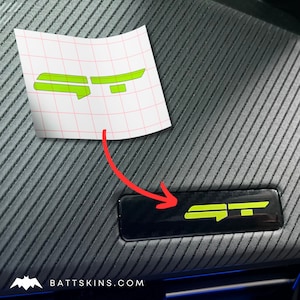 Puede incluir: Una moldura interior de coche de plástico negro con una pegatina de logotipo "GT" verde aplicada. La pegatina está en una hoja de transferencia transparente.