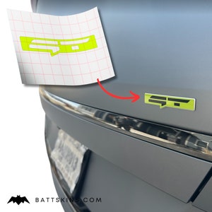 Puede incluir: Una pegatina de emblema "GT" verde y negro en un coche gris. La pegatina se aplica en la parte trasera del coche, por encima de la matrícula.