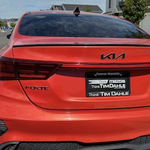 May include: Rear view of a bright red Kia Forte. The car has black accents, including the rear spoiler and a black mesh lower bumper. The Kia logo is visible, along with the word "FORTE" and a license plate that reads "Think Tim Dahle."