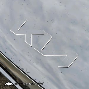 May include: Clear, transparent overlay featuring the Kia logo, a stylized emblem with interconnected geometric shapes. The logo is set against a dark, reflective surface. The website address BATTSKINS.COM is visible at the bottom.
