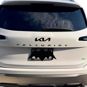 May include: Rear view of a white Kia Telluride SUV. The Kia logo and the word "Telluride" are in black lettering. A license plate with a bat-shaped design is visible. The vehicle has a rear window wiper.