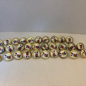 Puede incluir: Un conjunto de tiradores decorativos para armarios con un diseño floral. Cada tirador presenta un centro blanco con un patrón de flores rojas y azules, rodeado por un borde de metal dorado. Los tiradores están dispuestos sobre una superficie clara.