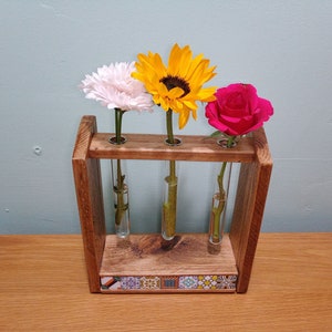 May include: A wooden flower vase with three glass test tubes for holding flowers. The vase is decorated with colourful tiles on the bottom. The vase is holding a white flower, a yellow sunflower, and a pink rose.