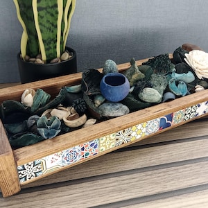 Könnte beinhalten: Ein rechteckiges Holztablett gefüllt mit getrocknetem Potpourri, darunter blaue, grüne und beige Elemente. Der Rand des Tabletts ist mit bunten, gemusterten Fliesen verziert. Eine kleine blaue Schale befindet sich inmitten des Potpourris. Eine Topf-Schlangenpflanze ist im Hintergrund.