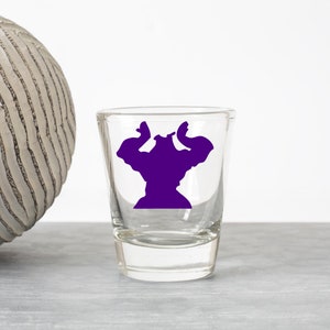 May include: A clear glass shot glass with a purple silhouette of a muscular figure flexing its biceps.
