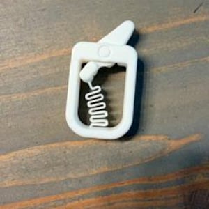 May include: A white plastic clip featuring a rectangular frame with a hinged lever at the top and a zigzag-shaped gripping mechanism inside the opening, designed to secure items firmly within its structure.