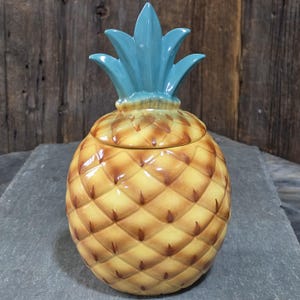 Puede incluir: Un tarro de cerámica con forma de piña y tapa extraíble. El cuerpo del tarro presenta una textura de patrón de diamantes en tonos amarillos y marrones, mientras que la tapa está rematada con una corona de hojas de color azul cerceta.