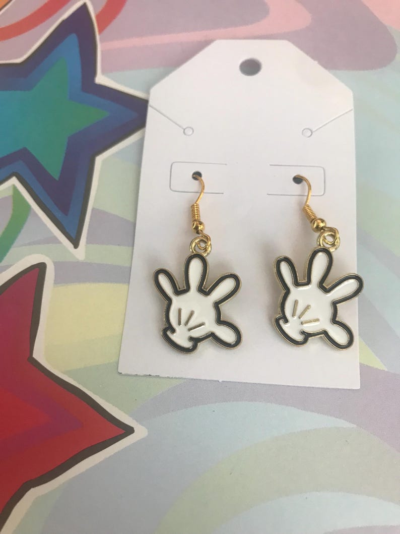 May include: A pair of gold dangle earrings with white Mickey Mouse glove charms. The gloves have black outlines and a single black line across the palm.