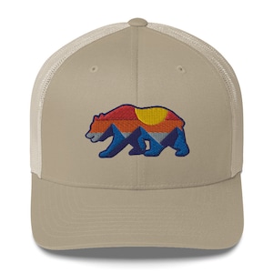 Puede incluir: Una gorra de camionero beige y blanca con una silueta de oso marrón bordada en la parte delantera. El oso está relleno con un diseño de cordillera de colores, que incluye rojo, naranja, amarillo, azul y verde.