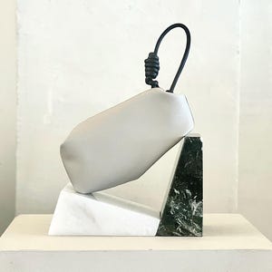 Könnte beinhalten: Eine hellgraue Ledertasche mit schwarzem Griff und Knotendetail. Die Tasche ruht auf einer geometrischen Skulptur aus weißem und grünem Marmor. Die Skulptur steht auf einer weißen Oberfläche.