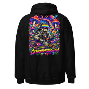 Könnte beinhalten: Ein schwarzer Kapuzenpullover mit einer lebendigen, mehrfarbigen Grafik auf der Rückseite. Das Design zeigt einen Astronauten mit regenbogenfarbener Brille und Kopfhörern an einem DJ-Pult, umgeben von bunten Pilzen, Kristallen und Planeten. Der Text CATSONDRUGS.COM ist in einer fetten, stilisierten Schrift am unteren Rand der Grafik gedruckt.