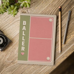 以下が含まれることがあります： セージグリーンの背表紙に白いブロック体でBALLERの文字と小さなピンクのハートが描かれた長方形のノート。表紙はダスティピンクで、2つの長方形のセクションと角に2つの白い円形のアクセントが配置されています。